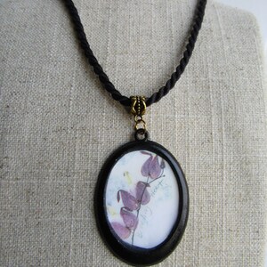 May include: A black cord necklace with an oval pendant featuring a pressed flower design. The pendant has a black frame and a white background with the text "Bleeding Heart".