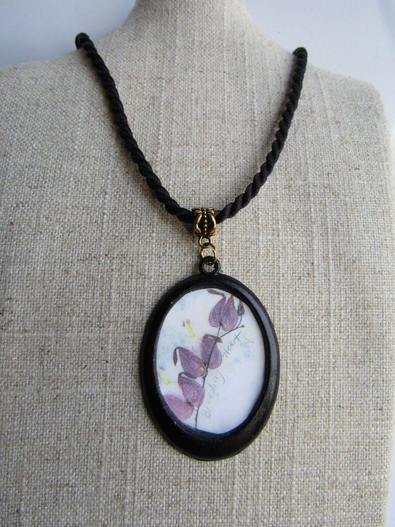 May include: A black cord necklace with an oval pendant featuring a pressed flower design. The pendant has a black frame and a white background with the text "Bleeding Heart".
