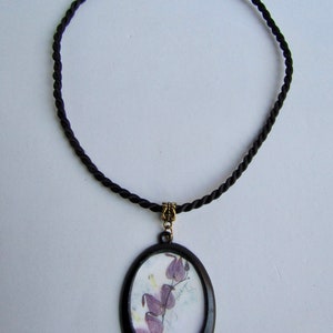 May include: A black cord necklace with a round pendant featuring a pressed flower design. The pendant has a black frame and a clear glass insert. The flower is purple and has the text "Bleeding Heart" written on the paper.