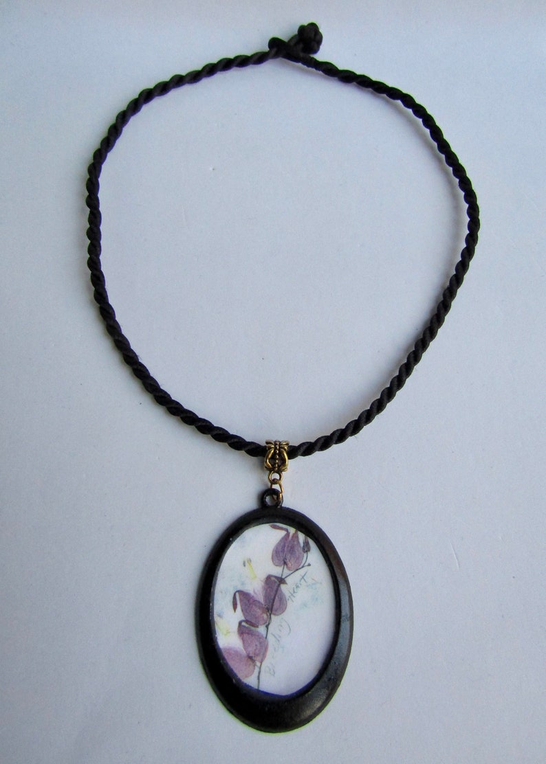 May include: A black cord necklace with a round pendant featuring a pressed flower design. The pendant has a black frame and a clear glass insert. The flower is purple and has the text "Bleeding Heart" written on the paper.