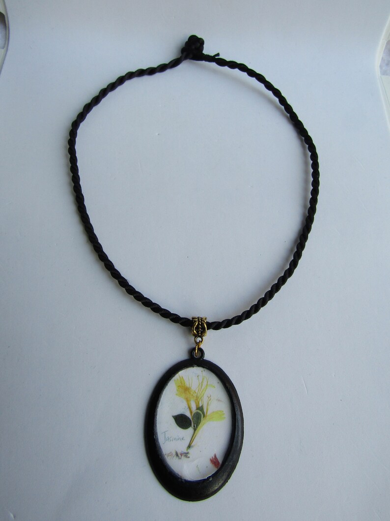Op de afbeelding: Zwarte koordketting met een ovale hanger. De hanger heeft een geperste bloem met het woord "Jasmine" eronder geschreven. De hanger is zwart omlijst met een goudkleurige balein.