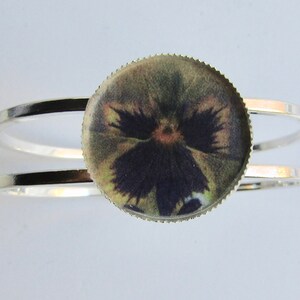 Op de afbeelding: Een zilveren armband met een ronde, heldere harscabochon met een geperste viooltjesbloem in paarse en groene tinten.
