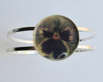 May include: A silver bangle bracelet with a round, clear resin cabochon featuring a pressed pansy flower with purple and green hues.