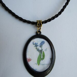 Puede incluir: Un collar de cord&oacute;n negro con un cierre dorado y un colgante ovalado negro con una flor de nomeolvides prensada con el texto "Forget-me-nots".