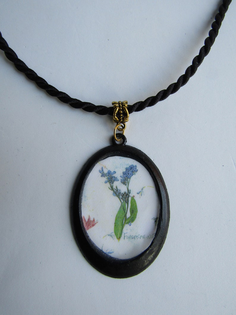 Puede incluir: Un collar de cord&oacute;n negro con un cierre dorado y un colgante ovalado negro con una flor de nomeolvides prensada con el texto "Forget-me-nots".