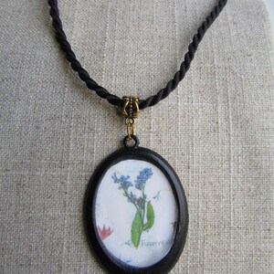 Puede incluir: Un collar de cord&oacute;n negro con un colgante ovalado negro que presenta una flor de nomeolvides prensada con el texto "Forget-me-not" escrito en el papel.