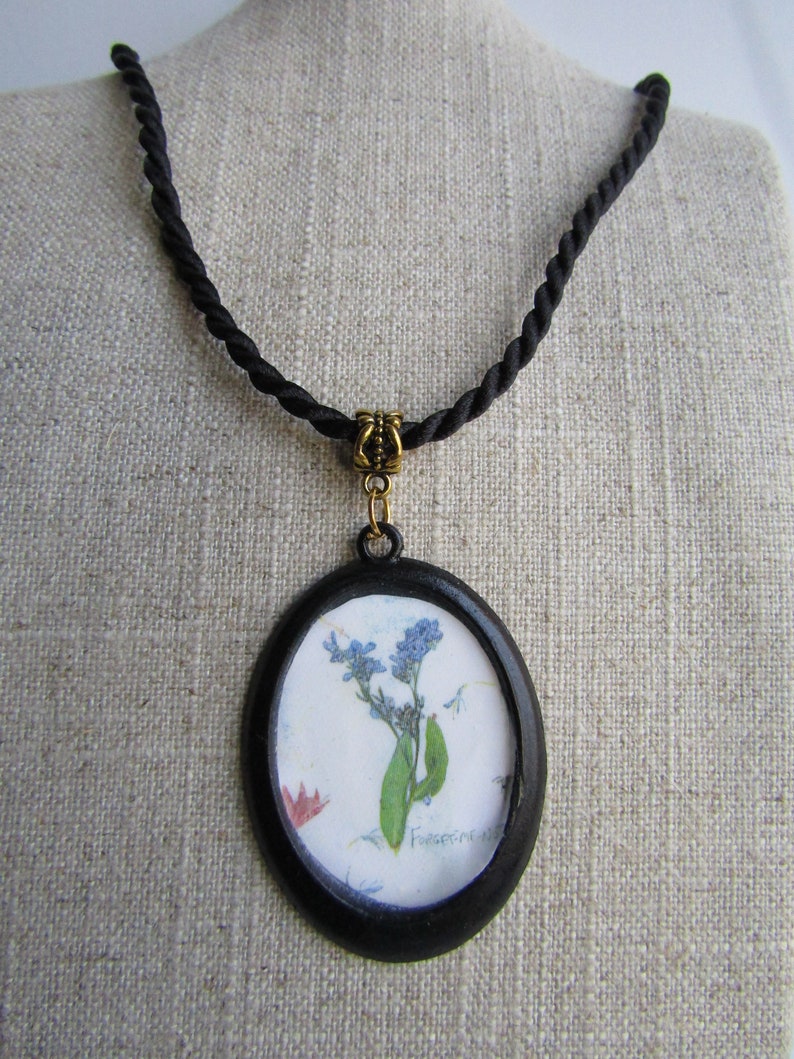Puede incluir: Un collar de cord&oacute;n negro con un colgante ovalado negro que presenta una flor de nomeolvides prensada con el texto "Forget-me-not" escrito en el papel.