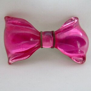 Könnte beinhalten: Eine Brosche in Form einer Schleife aus rosa Glas mit einem goldenen Metallzentrum.
