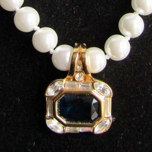 Op de afbeelding: Een gouden ketting met een hanger met een donkerblauwe edelsteen omringd door heldere strass steentjes. De ketting is gemaakt van witte parels.