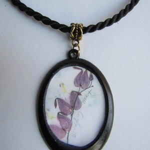 May include: A black oval pendant necklace with a black cord. The pendant features a pressed flower image of a bleeding heart flower with the text "Bleeding Heart" written on the paper.