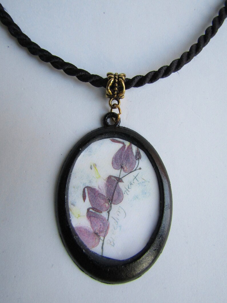 May include: A black oval pendant necklace with a black cord. The pendant features a pressed flower image of a bleeding heart flower with the text "Bleeding Heart" written on the paper.