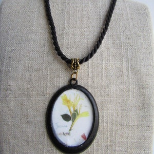 Op de afbeelding: Een zwarte ovale hangerketting met een geperste gele bloem met het woord "Jasmine" op de afbeelding. De hanger hangt aan een zwart touwketting met een goudkleurige sluiting.