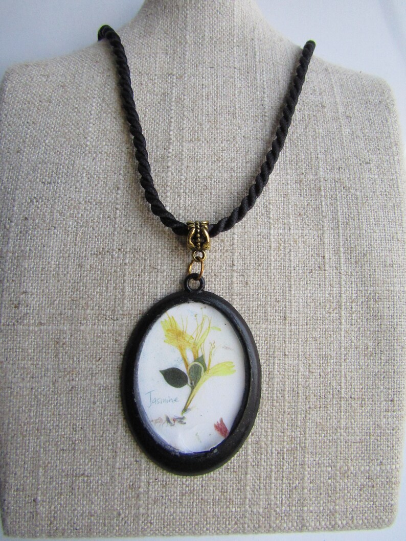 Op de afbeelding: Een zwarte ovale hangerketting met een geperste gele bloem met het woord "Jasmine" op de afbeelding. De hanger hangt aan een zwart touwketting met een goudkleurige sluiting.