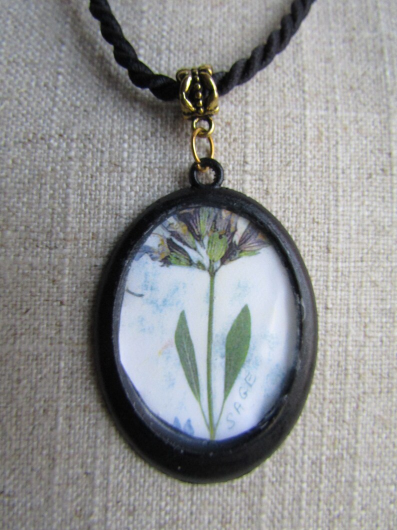 May include: A black oval pendant necklace featuring a pressed flower design. The flower is a sage plant with a white background and the word "SAGE" printed in the bottom right corner.