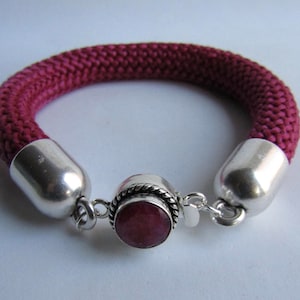 Peut inclure: Un bracelet en corde tressée marron avec des embouts argentés et un fermoir argenté orné d'une pierre rouge.