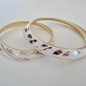 May include: Two white bangle bracelets with brown swirl patterns. The bracelets are made of a smooth, shiny material.