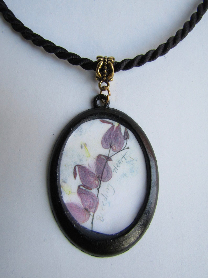 May include: A black oval pendant necklace with a black cord. The pendant features a pressed flower design of a bleeding heart flower with the text "Bleeding Heart" written on the paper.