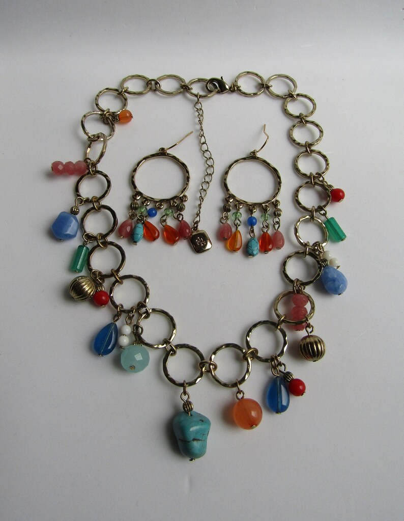 May include: A gold chain necklace with multiple colorful beads and charms, including turquoise, coral, and blue glass. The necklace is paired with matching earrings with similar charms.