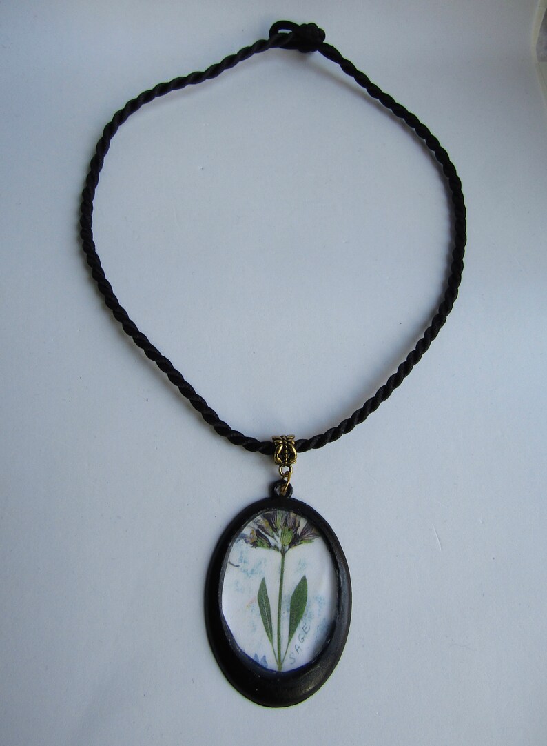 May include: A black cord necklace with a round pendant featuring a pressed sage flower inside a black frame. The pendant has the word "SAGE" written on it.