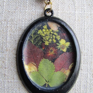 Op de afbeelding: Een zwarte ovale hangerketting met een gouden sluiting. De hanger bevat een geperst bloemenmotief met groene, rode en gele bloemen.
