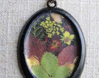 May include: A black oval pendant necklace with a gold clasp. The pendant contains a pressed flower design with green, red, and yellow flowers.