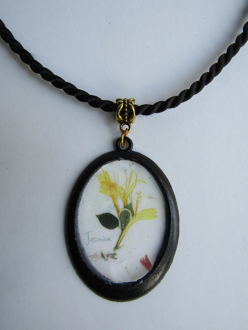 Op de afbeelding: Een zwarte koordketting met een goudkleurige sluiting en een zwarte ovale hanger. De hanger toont een geperste gele bloem met het woord "Jasmine" eronder.