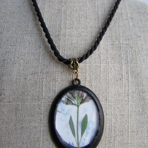 May include: A black cord necklace with a round pendant featuring a pressed flower and the word "SAGE" in white lettering.