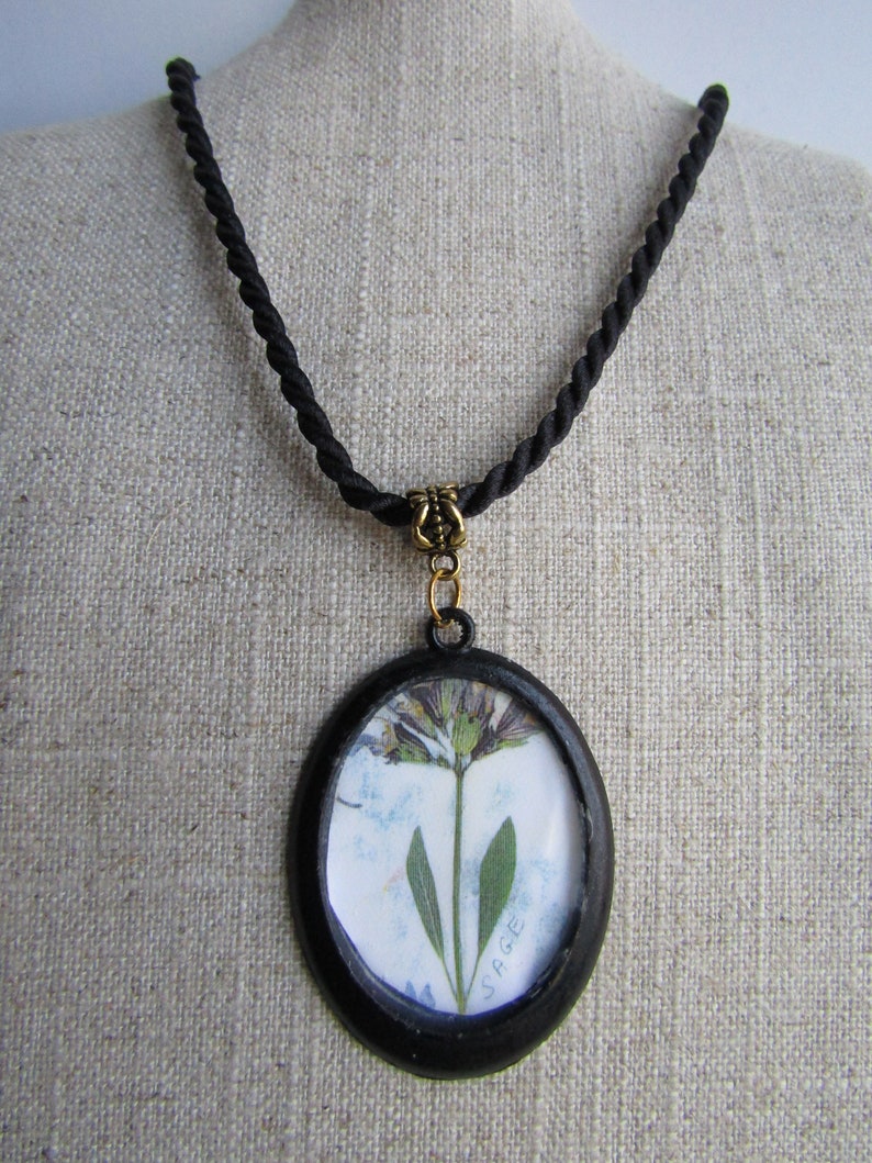 May include: A black cord necklace with a round pendant featuring a pressed flower and the word "SAGE" in white lettering.