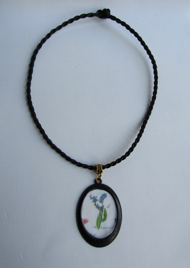 Puede incluir: Un collar de cord&oacute;n negro con un colgante ovalado negro que presenta un dise&ntilde;o de flores prensadas. La flor es azul y blanca con la palabra "No me olvides" escrita en cursiva.