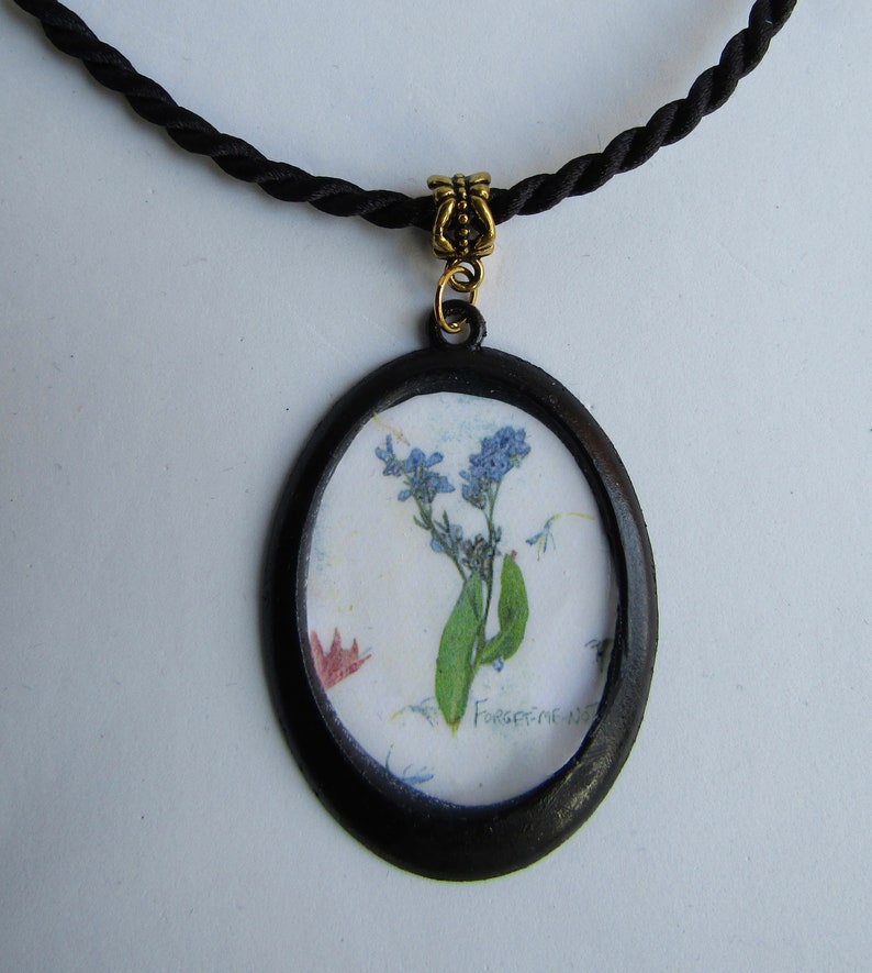 Puede incluir: Un collar con un colgante ovalado negro que contiene una flor de nomeolvides prensada. El colgante est&aacute; en un cord&oacute;n negro.
