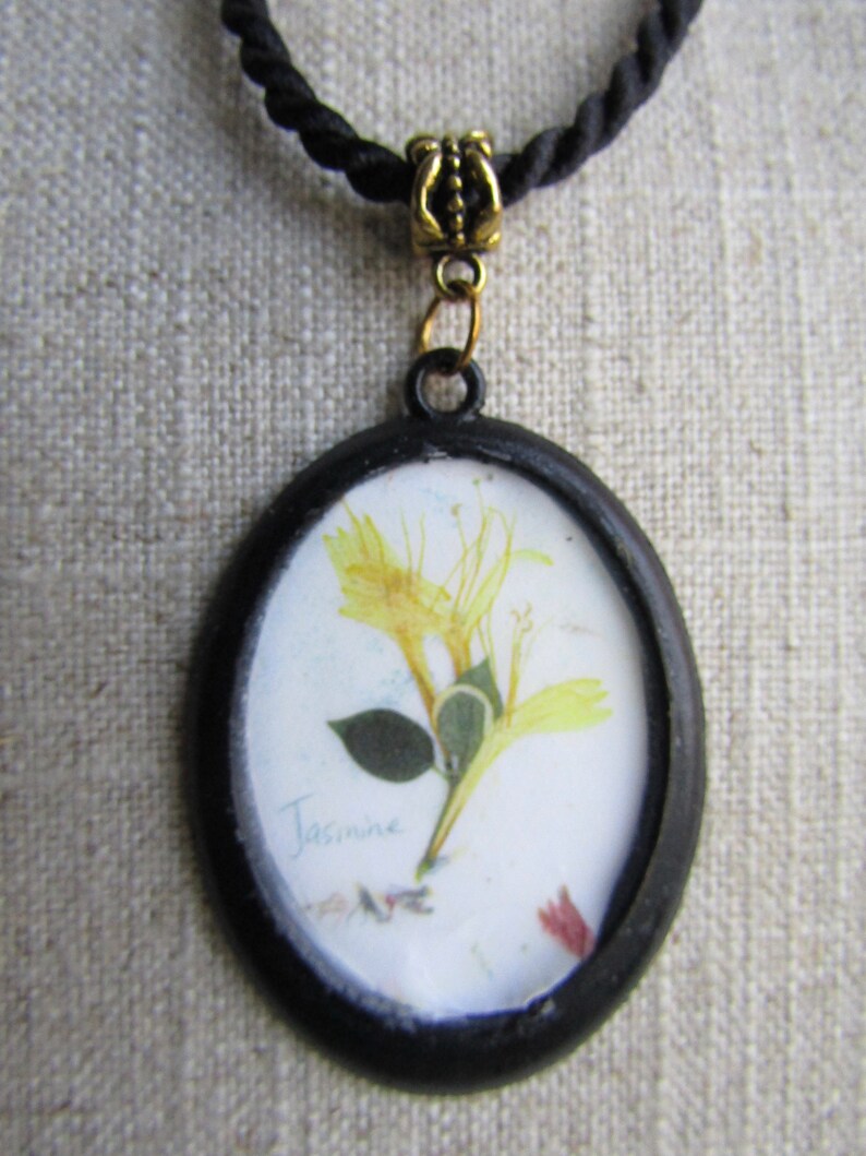 Op de afbeelding: Ovale hangerketting met een geperste gele bloem en het woord "Jasmine" op een witte achtergrond. De hanger heeft een zwart frame en hangt aan een zwarte koordketting met een goudkleurige sluiting.