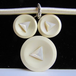 May include: A white necklace featuring three white buttons, each with a white triangle in the centre. The buttons are attached to a white satin ribbon.