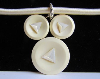 May include: A white necklace with three white buttons, each with a white triangle in the center. The buttons are attached to a white satin ribbon.
