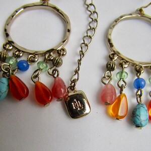 May include: Gold hoop earrings with a dangling charm featuring a variety of colorful beads, including turquoise, orange, pink, and blue. The charm also includes a small gold tag with the letters "RL" engraved on it.