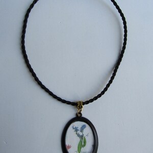 Puede incluir: Un collar de cord&oacute;n negro con un colgante ovalado negro que presenta un dise&ntilde;o de flores prensadas. La flor es azul y blanca con la palabra "No me olvides" escrita en cursiva.