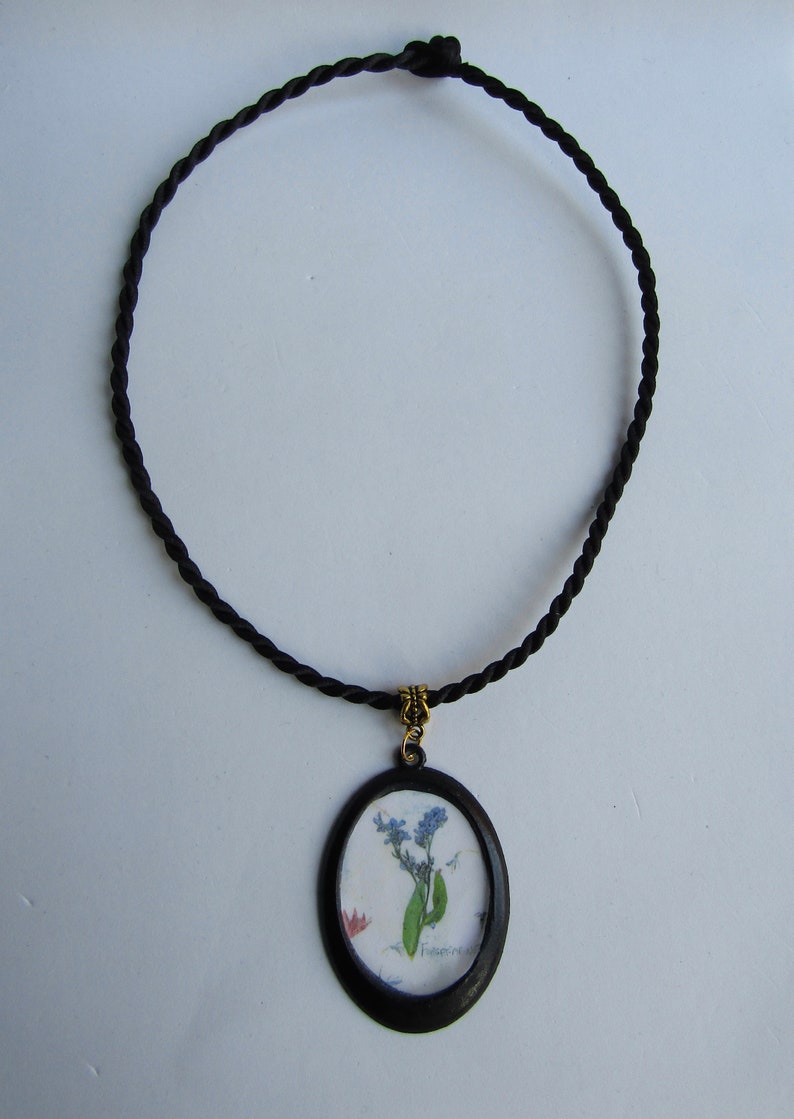 Puede incluir: Un collar de cord&oacute;n negro con un colgante ovalado negro que presenta un dise&ntilde;o de flores prensadas. La flor es azul y blanca con la palabra "No me olvides" escrita en cursiva.