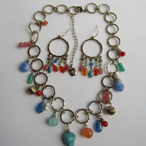 May include: A gold chain necklace with multiple colorful gemstone charms and matching earrings. The necklace features a series of connected rings with dangling charms, including turquoise, red, orange, and blue gemstones.