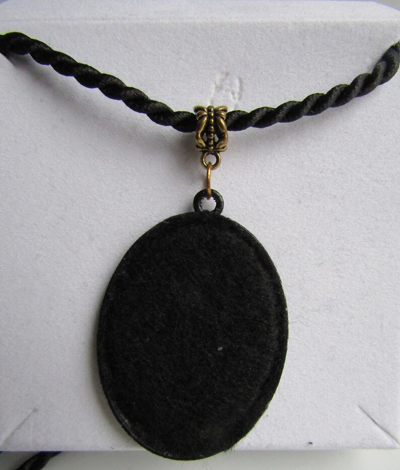 Puede incluir: Un collar con un colgante ovalado negro y un cierre dorado. El colgante est&aacute; hecho de un material negro liso y cuelga de un cord&oacute;n negro.