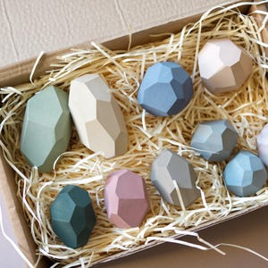 Puede incluir: Un conjunto de diez formas geométricas de madera en varios tonos de verde, azul, rosa y marrón. Las formas están dispuestas en una caja llena de papel triturado.