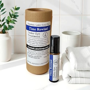 May include: A brown cardboard tube with the text "Crystal Lake Soap Co. Time Rewind Under Eye Treatment" and a small bottle of "Time Rewind Under Eye Treatment" on a white tiled surface. The bottle is clear with a brown cap and a label with blue and white text.