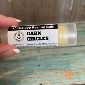 May include: A white tube with a black label that reads "Under Eye Rescue Balm" and "DARK CIRCLES". The label also lists the ingredients: Cocoa & Shea Butters, Coconut, Avocado Oils, Beeswax, Rosehip, Carrot.