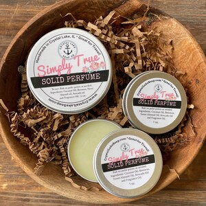 May include: Two small silver tins with white lids containing solid perfume. The tins are labeled "Simply True Solid Perfume" and list the ingredients: Coconut Oil, Beeswax, Sweet Almond Oil, Avocado Oil, and Fragrance Oil. The tins are sitting in a brown paper shred basket.