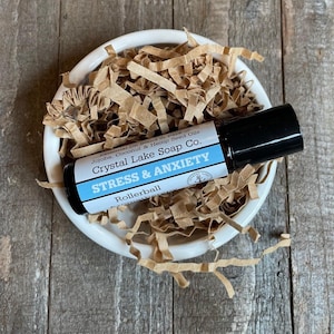 May include: A rollerball bottle labeled "STRESS & ANXIETY" by Crystal Lake Soap Co. rests in a white bowl filled with brown paper shreds. The bottle has a black cap and a blue and white label. The bowl sits on a wooden surface.
