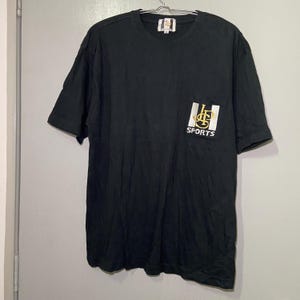 May include: Black short-sleeved t-shirt with a white pocket featuring a gold logo and the text "SPORTS".