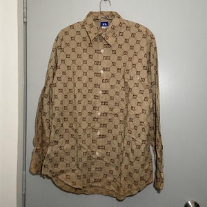 Puede incluir: Camisa abotonada de color beige con un patrón geométrico marrón repetido. La camisa de manga larga está colgada de una percha de metal. La camisa tiene cuello y puños abotonados.