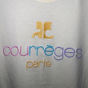 May include: White t-shirt with a gold embroidered logo that says "Courr&egrave;ges Paris". The logo is in a stylised font with the "C" in the logo being a stylised "C" with a curved line.