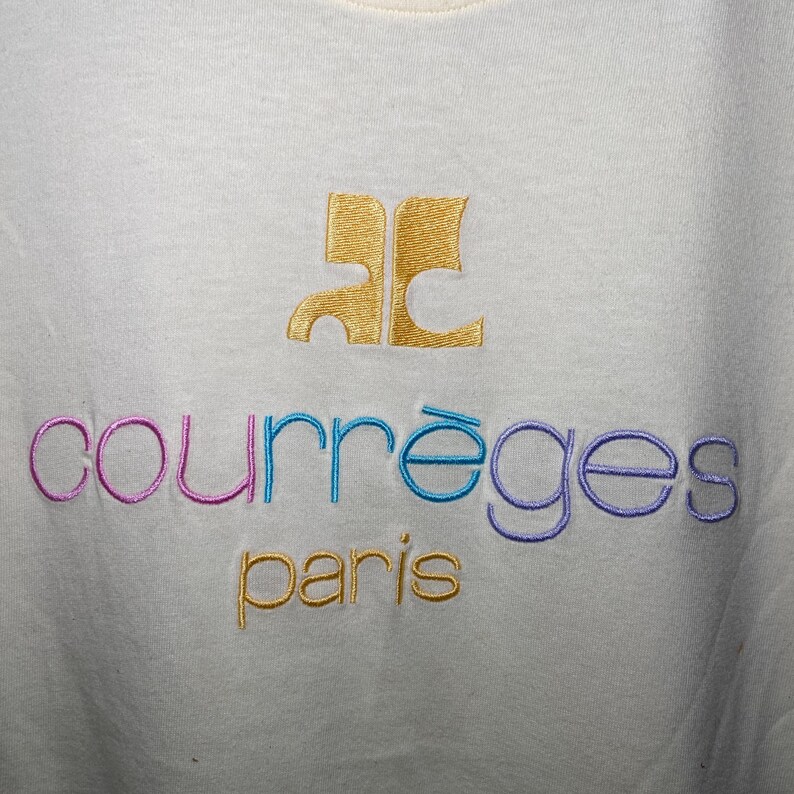 May include: White t-shirt with a gold embroidered logo that says "Courr&egrave;ges Paris". The logo is in a stylised font with the "C" in the logo being a stylised "C" with a curved line.