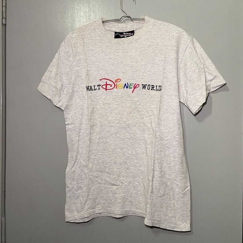Pu&ograve; includere: Una maglietta grigia con un logo Walt Disney World ricamato a colori.