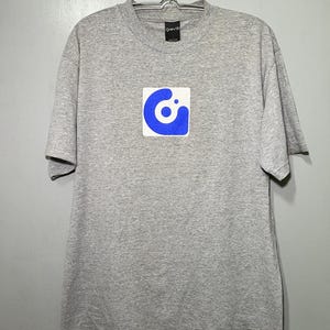 May include: A gray t-shirt with a blue logo of a circle with a smaller circle inside, all within a square. The logo says "Gravis" in white text.