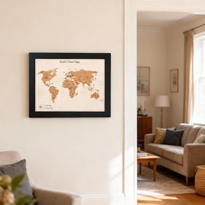 May include: A framed cork world map with a black frame. The map displays continents in brown tones, with pins indicating locations. The text "Noel's Travel Map" is at the top. The map is mounted on a cream-coloured wall.